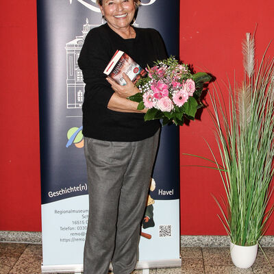 Elisabeth Herrmann mit ihrem Buch und einem Strauß Blumen in der Hand lächelt in die Kamera.