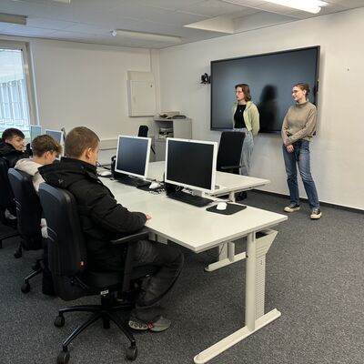 Einige Schülerinnen und Schüler sitzen an PCs und hören den Ausbildungsleiterinnen zu.